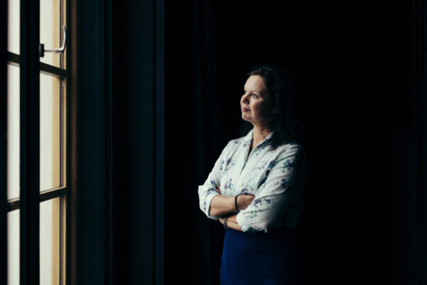 Porträtt av Ann-Luise Bertell mot mörk bakgrund som tittar ut genom fönstret